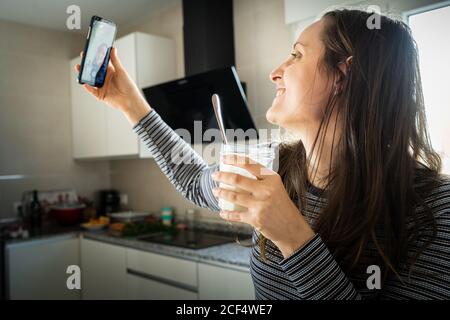 Junge Frau mit einem Glas gesunden Joghurts lächelt und posiert für Selfie in der Küche Stockfoto
