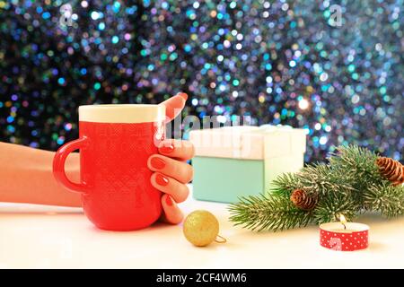 Frau Hand hält eine Tasse Kaffee auf einem weihnachtlichen Hintergrund. Weihnachtsgeschenkschachteln und grüner Tannenbaum über leuchtendem Tisch. Stockfoto
