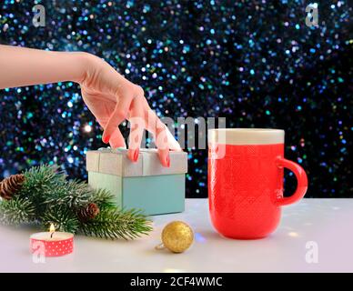 Die Hand der Frau hält eine Geschenkbox auf einem weihnachtlichen Hintergrund. Rote Tasse Kaffee und grüne Fichte über einem glänzenden Tisch. Stockfoto