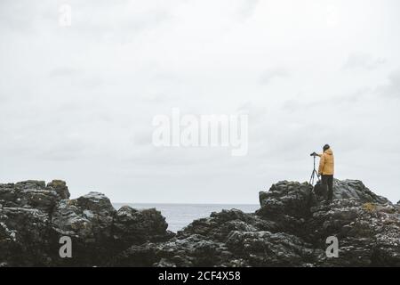 Rückansicht des männlichen Reisenden, der auf Felsen mit Kamera steht Auf Stativ und Aufnahme von Seestück auf trüb düster Tag an der nordirischen Küste Stockfoto