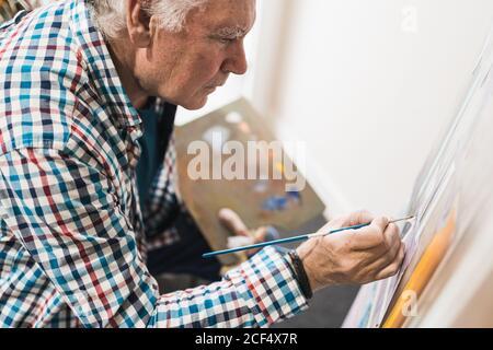 Seitenansicht von Crop Senior männlichen Künstler in legerer Kleidung Zeichnung Bild mit Pinsel auf Papier auf Staffelei halten Palette Mit Farben in der kreativen Kunstwerkstatt Stockfoto
