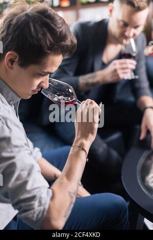 Sommelier untersucht Wein. Zugeschnittenes Bild eines selbstbewussten männlichen Sommeliers, der Wein untersucht, während er ihn riecht und sich an den Holztisch mit Weinregal i lehnt Stockfoto