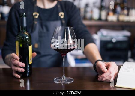 Sommelier untersucht Wein. Zugeschnittenes Bild eines selbstbewussten männlichen Sommeliers, der Wein untersucht, während er ihn riecht und sich an den Holztisch mit Weinregal i lehnt Stockfoto