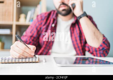 Ein anonymer Mann schreibt in einem Notizbuch. Der Mann arbeitet von zu Hause aus. Der Unternehmer spricht auf dem Handy Stockfoto