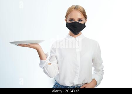 Kellnerin in Maske mit Teller in der Hand auf Seite isoliert Auf weißem Studiohintergrund Stockfoto