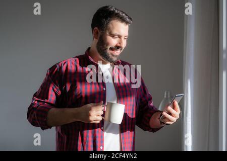 Ein junger Mann schaut auf das Handy. Der Mann arbeitet von zu Hause aus. Der Unternehmer hält ein Telefon und trinkt aus einem Becher. Er lächelt Stockfoto