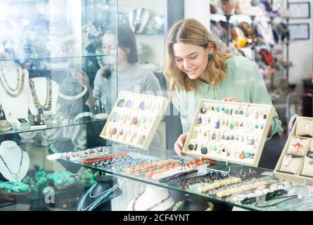 Junge attraktive Verkäuferin, die hinter dem Fenster des Juweliershops arbeitet und verschiedene Halsketten, Armbänder, Anhänger und Ohrringe aus Halbedelei arrangiert Stockfoto