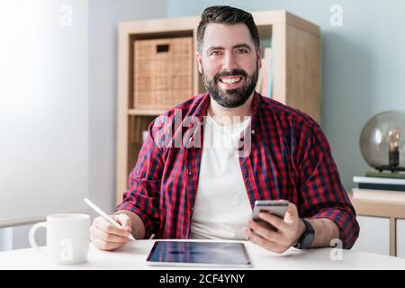 Ein junger Mann schreibt auf einem Tablet. Der Mann arbeitet von zu Hause aus. Der Unternehmer hält ein Telefon Stockfoto