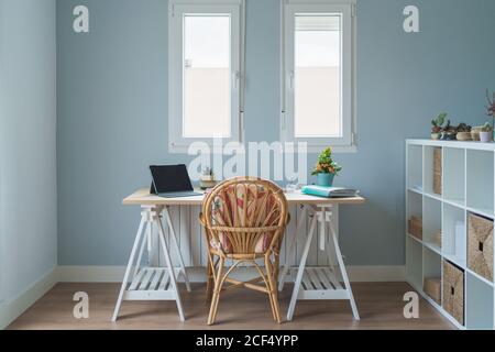 Interieur des modernen Zimmer und Arbeitsplatz mit Holzschreibtisch mit Laptop und Ordner und whicker Stuhl in der Nähe von Holzregalen mit Kisten und Topfpflanzen Stockfoto