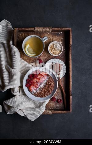Blick von oben auf gesundes hausgemachtes Frühstück mit Müsli und Beeren Smoothie Schüssel und Tasse Kräutertee mit Zitrone serviert Auf Holztablett auf schwarzem Hintergrund Stockfoto