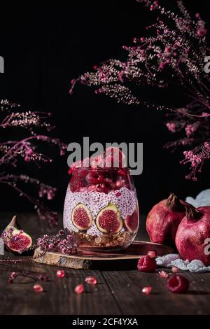 Chia Pudding mit frischen Feigen und Himbeeren einschließlich Granatapfel und Creme im Glas am Tisch zwischen rosa Blütenbünden auf Schwarzer Hintergrund Stockfoto