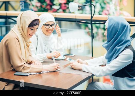 Drei hübsche muslimische Frauen mit Hijab im Café. Sitzen auf Sofas an einem Tisch und reden. Ein Mädchen mit Brille Stockfoto