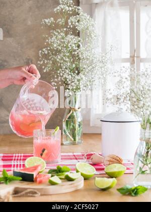 Eisrosa Getränk mit Wassermelone und Limettenscheiben in Gießen Glas aus kaltem Glas auf rustikalem Küchentisch nebenan Bündel von Gypsophila Blumen bei Tageslicht Stockfoto