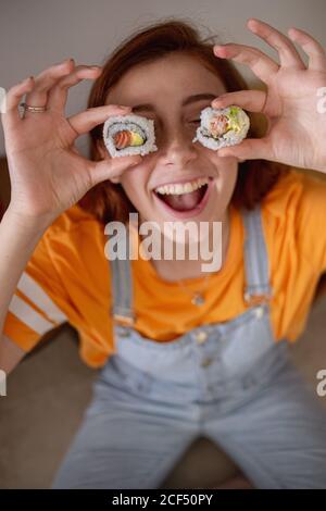 Von oben entzückten junge Frau, die schmackhaftes Sushi in der Nähe der Augen hielt Beim Mittagessen zu Hause Stockfoto
