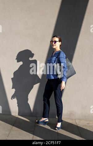 Ganzkörper weibliche Manager in Sonnenbrille und elegantes Outfit lächelnd Und auf die Kamera schauen, während man gegen die graue Gebäudewand steht An der City Street Stockfoto