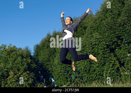 Niedriger Winkel der barfuß weibliche Manager in eleganten Outfit heben Arme und springen gegen grüne Bäume und wolkenlosen Himmel während Erfolg im Park feiern Stockfoto