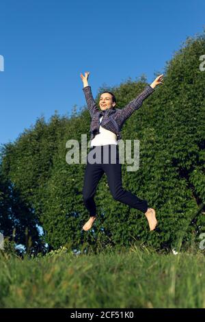 Niedriger Winkel der barfuß weibliche Manager in eleganten Outfit heben Arme und springen gegen grüne Bäume und wolkenlosen Himmel während Erfolg im Park feiern Stockfoto