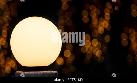 Runde weiße Straßenlaterne leuchtet mit warmem Leuchten, im Hintergrund Bokeh diffuse Kreise aus Girlanden in der Nacht auf dunklem Hintergrund, Urlaubspostkarte mit Co Stockfoto