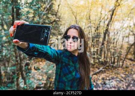 Frau im karierten Hemd nimmt Selfie auf Handy, während Auf Waldholz stehend Stockfoto