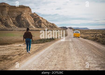 Rückansicht einer nicht erkennbaren Dame im Urlaub in stilvoller Freizeitkleidung, die auf einer sandigen Straße zwischen verlassenen Gelände und Bergen in Bardenas Reales, Navarra, Spanien, spazieren geht Stockfoto