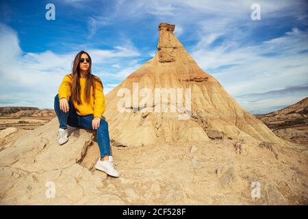 Fröhliche junge weibliche Reisende in stilvollen Casual tragen sitzen in braunen Hügel mit blauem Himmel auf Hintergrund in Bardenas Reales, Navarra, Spanien Stockfoto