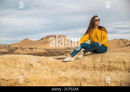 Fröhliche junge weibliche Reisende in stilvollen Casual tragen sitzen in braunen Hügel mit blauem Himmel auf Hintergrund in Bardenas Reales, Navarra, Spanien Stockfoto