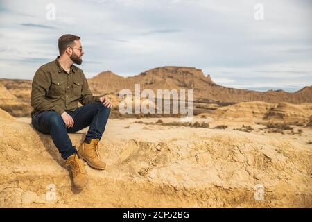 Niedriger Winkel des Inhalts bärtige männlich in Freizeitkleidung und Sonnenbrille lächelnd, während sie am Fuß des braunen Berges mit ruht Blauer Himmel im Hintergrund bei Bardenas Reales Stockfoto