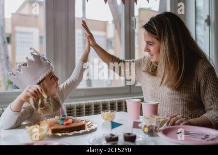 Nettes kleines Mädchen in legerer Kleidung und Papier Krone geben Hohe fünf mit Mutter, während sie am Tisch sitzt und isst Leckeres Essen im Urlaub Stockfoto