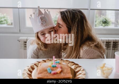 Liebevolle Mutter küssen und gratulieren kleinen Mädchen zusammen, während Ausgaben Zeit während der Geburtstagsfeier am Tisch zu Hause Stockfoto