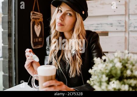 Langhaarige trendige schöne blonde Frau, die aus einem Glas köstlichen schäumenden Kaffees trinkt und die Kamera anschaut Stockfoto