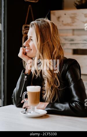 Seitenansicht der langhaarigen, trendigen, schönen blonden Frau, die in einem Café-Geschäft sitzt und von einem Glas köstlichen schäumenden Kaffees trinkt Stockfoto