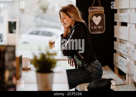 Seitenansicht einer langhaarigen, trendigen, schönen blonden Frau, die in einem Café-Geschäft sitzt und von einem Glas köstlichen schäumenden Kaffees mit geschlossenen Augen trinkt Stockfoto