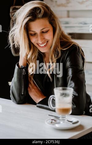 Seitenansicht einer langhaarigen, trendigen, schönen blonden Frau, die in einem Café-Geschäft sitzt und von einem Glas köstlichen schäumenden Kaffees mit geschlossenen Augen trinkt Stockfoto