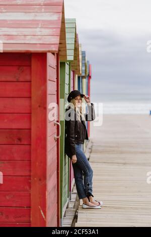 Trendige langhaarige blonde Frau in schwarzer Mütze und Lederjacke lächelt hell vor der Kamera und lehnt sich an die Wand der hölzernen Strandhütten Stockfoto