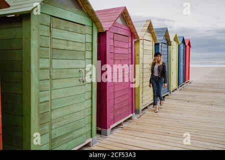 Trendige langhaarige blonde Frau in schwarzer Mütze und Lederjacke lächelt hell an der Kamera und an der hölzernen Strandhütte vorbei Stockfoto