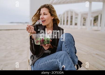 Stilvolle langhaarige blonde Frau in schwarzer Jacke genießt gesunden grünen Salat, während sie lächelnd auf der Holzterrasse an der Küste sitzt und wegschaut Stockfoto