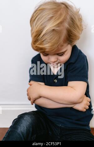 Genervt blonde kleinen Jungen in lässigen Kleidung sitzt auf der Boden mit gekreuzten Armen Stockfoto