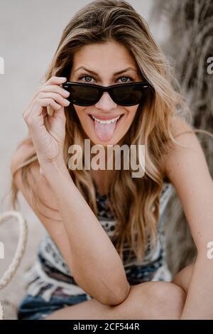 Nahaufnahme eines Porträts einer hübschen blonden Frau in schwarzer Sonnenbrille, die auf Sand sitzt und die Kamera anschaut und aus der Zunge ragt Stockfoto