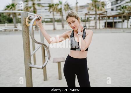 Motivierte, sportliche Frau in aktiver Kleidung, die mit einer Metallstange steht, während sie sich an einem Sandstrand ausruhen und die Kamera betrachten kann Stockfoto