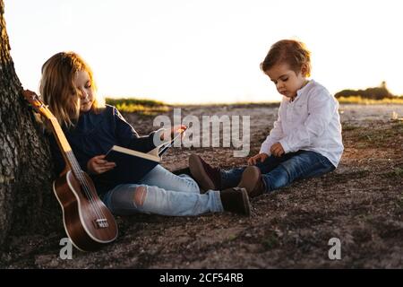 Seitenansicht von liebenswert kleines Mädchen lesen interessante Geschichte zu Jüngerer Bruder, während er zusammen unter dem Baum mit Ukulele-Gitarre sitzt Im Sommer Tag auf dem Land Stockfoto