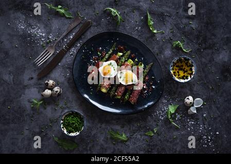 Von oben leckerer Spargel mit gebratenen Wachteleiern und Kräutern Platziert auf einer Schüssel neben Besteck auf Marmor-Tischplatte Stockfoto