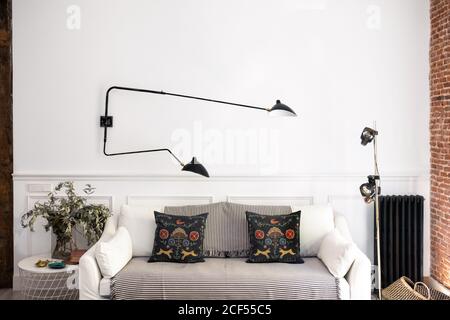 Bequemes Sofa mit weichen Kissen unter moderner Metalllampe Neben Tischen mit Dekorationen in stilvollen gemütlichen Wohnzimmer an Zu Hause Stockfoto