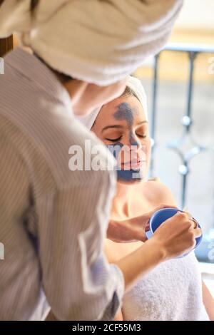 Anonyme Frau, die während des Eingriffs zu Hause eine blaue Tonmaske auf das Gesicht einer ruhigen Frau mit geschlossenen Augen in einem weißen Handtuch anwendet Stockfoto