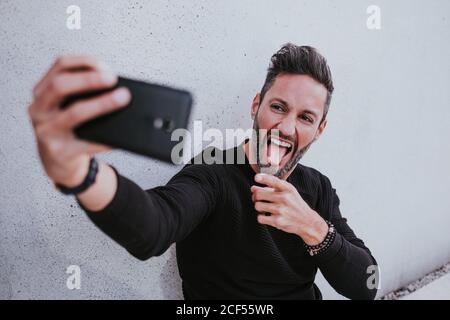 Mittelalter gut aussehend elegant glücklich männlich in Casual tragen Einnahme Selfie auf Handy und sitzen in der Nähe der grauen Wand Stockfoto