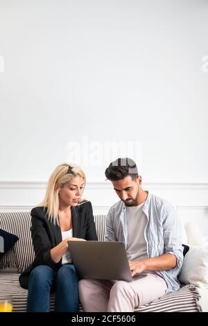Blonde Frau und ein gutaussehender Mann mit ethnischem Bart sitzen in legerer Kleidung auf dem Sofa und stöbern gemeinsam in einem Laptop zu Hause Stockfoto