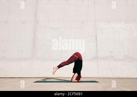 Seitenansicht einer nicht erkennbaren barfüßigen Sportlerin im aktiven Stehen Kopf nach unten in sirsasana Position mit Beinen angehoben auf Sport Mattentraining allein auf der Straße gegen Betonwand am Tag Stockfoto
