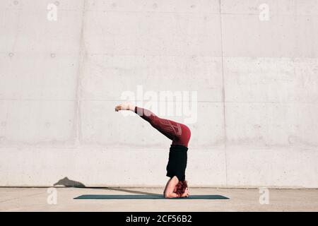 Seitenansicht einer nicht erkennbaren barfüßigen Sportlerin im aktiven Stehen Kopf nach unten in sirsasana Position mit Beinen angehoben auf Sport Mattentraining allein auf der Straße gegen Betonwand am Tag Stockfoto