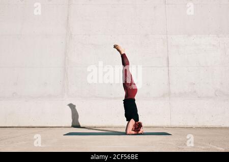Seitenansicht einer nicht erkennbaren barfüßigen Sportlerin im aktiven Stehen Kopf nach unten in sirsasana Position mit Beinen angehoben auf Sport Mattentraining allein auf der Straße gegen Betonwand am Tag Stockfoto