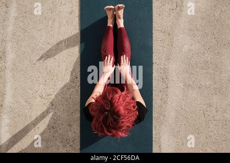 Von oben aus gesehen, wie eine anonyme barfuss-Frau in Sportkleidung nach dem Yoga-Training auf der Straße auf der Matte sitzt Stockfoto