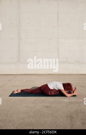Seitenansicht einer anonymen barfußigen Frau in Sportkleidung, die Yoga in Affenhaltung macht.nach vorne beugen, auf der Matte alleine auf der Straße gegen eine Betonwand trainieren Stockfoto